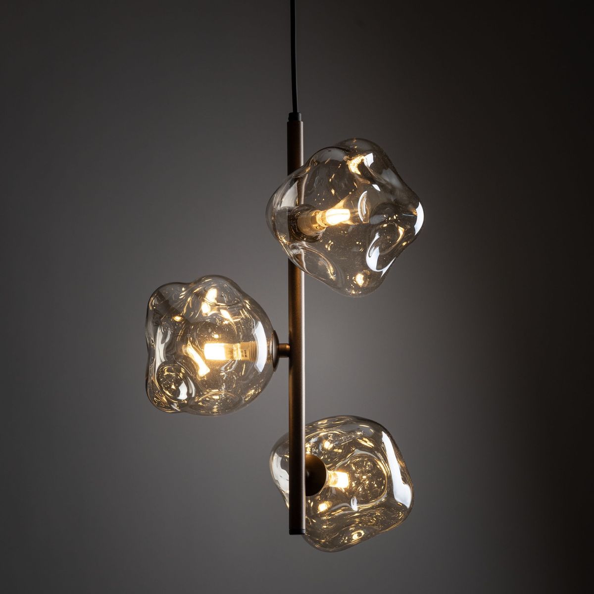 Brązowa lampa wisząca Lava Brown z trzema kloszami w kształcie nieregularnych brył, wykonana z przezroczystego szkła. Lampa umieszczona na poziomej metalowej rurze, zawieszona na czarnym przewodzie, w subtelnym oświetleniu.