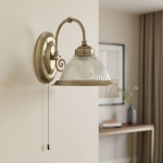 Kinkiet American Diner 1-punktowy w antycznym mosiądzu, z okrągłą podstawą i przezroczystym, żłobionym kloszem. Uchwyt w formie zakrętu, z widocznym włącznikiem na sznurku. Produkt zamontowany na jasnej ścianie, w tle z elementami aranżacyjnymi.