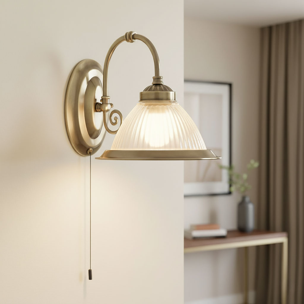 Kinkiet American Diner w antycznym mosiądzu, z przezroczystym, żłobionym kloszem. Zawieszony na ścianie, z ozdobnym uchwytem w kształcie spirali i przewodem do włączania. Tło jasne, z elementami dekoracyjnymi w aranżacji.