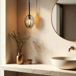 Czarna lampa wisząca Pearl Cognac z bursztynowym kloszem o owalnym kształcie, zawieszona na cienkim kablu, w eleganckim wnętrzu z lustrem i ceramiczną umywalką. W tle widoczny złoty wazon z suszonymi kwiatami.