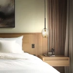Lampa wisząca Pearl Cognac z czarnym kablem i bursztynowym kloszem, zawieszona nad łóżkiem w nowoczesnej sypialni. Klosz ma owalny kształt, a w tle widoczne są drewniane panele i stół nocny z dekoracjami.