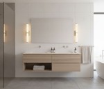 Kinkiet LED IP44 w kolorze złotym, jednostkowy, zamontowany na ścianie w łazience. Oświetlenie w formie prostokątnych lamp z kloszem z białego materiału, obok lustra nad umywalką. W tle drewniana szafka z ręcznikami i kosmetykami.