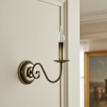 Asti kinkiet 1-punktowy w patynie, z pojedynczym ramieniem w kształcie spirali, zakończonym uchwytem na świecę. Kinkiet montowany na ścianie, z przezroczystą żarówką w kształcie płomienia. Tło w jasnym kolorze, z elementami dekoracyjnymi w zasięgu wzroku.