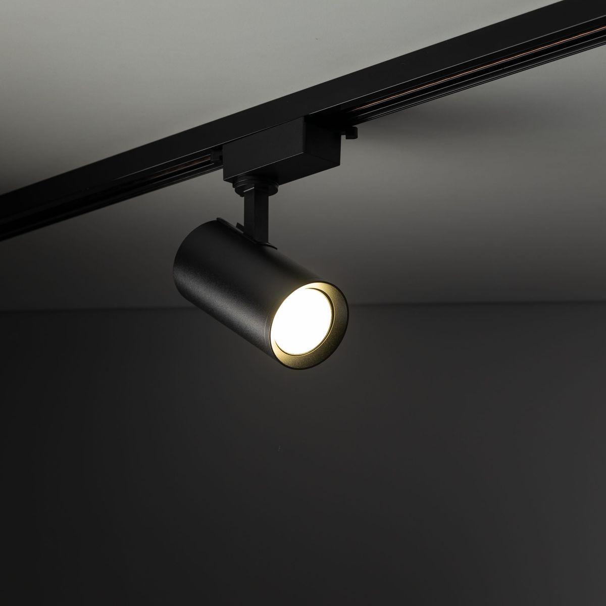 Reflektor spot Tracer 1xGU10 w kolorze czarnym, mocowany na szynie 1-fazowej. Widziany z boku, z włączonym światłem, z cylindrycznym korpusem i widoczną żarówką, umieszczony w minimalistycznym wnętrzu na tle szarym.
