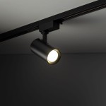 Reflektor spot Tracer 1xGU10 w kolorze czarnym, mocowany na szynie 1-fazowej. Widziany z boku, z włączonym światłem, z cylindrycznym korpusem i widoczną żarówką, umieszczony w minimalistycznym wnętrzu na tle szarym.