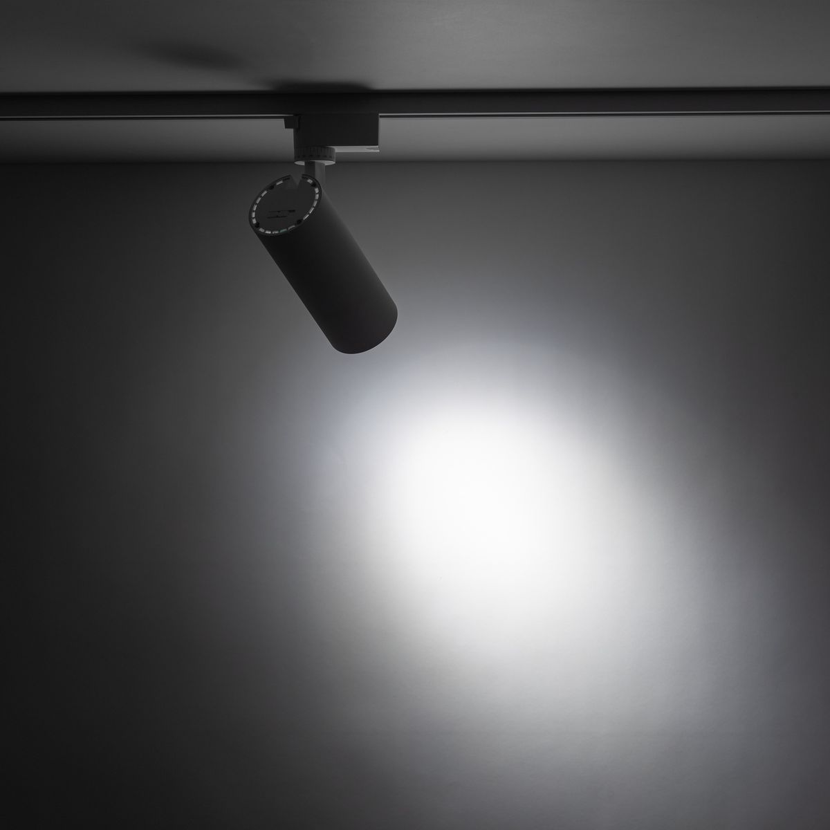 Reflektor Tracer 1xLED 20W CCT w kolorze białym, zamontowany na szynie 1-fazowej, w pozycji wiszącej. Cylindryczny kształt z widocznym źródłem światła, emitującym jasne światło na szare tło.