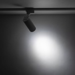 Reflektor Tracer 1xLED 20W CCT w kolorze białym, zamontowany na szynie 1-fazowej, w pozycji wiszącej. Cylindryczny kształt z widocznym źródłem światła, emitującym jasne światło na szare tło.