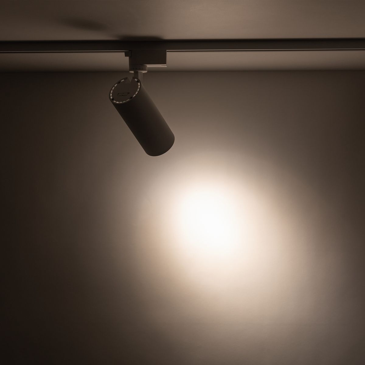 Reflektor Tracer spot 1xLED 20W CCT w kolorze białym, zamocowany na szynie 1-fazowej. Ujęcie z boku, pokazujące cylindryczny kształt lampy oraz emitowane światło kierujące się w dół na gładką powierzchnię.