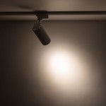 Reflektor Tracer spot 1xLED 20W CCT w kolorze białym, zamocowany na szynie 1-fazowej. Ujęcie z boku, pokazujące cylindryczny kształt lampy oraz emitowane światło kierujące się w dół na gładką powierzchnię.