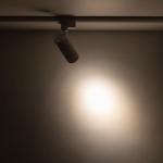 Reflektor spot Tracer 1xLED 10W CCT w kolorze białym, zamontowany na szynie 1-fazowej, skierowany w dół. Emituje światło na ścianę, tworząc okrągły, jasny punkt o miękkich krawędziach na tle ciemnej powierzchni.
