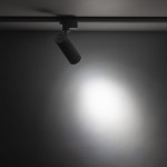 Reflektor spot Tracer 1xLED 10W CCT w białej obudowie, zamontowany na szynie 1-fazowej. Ujęcie z boku, skierowany w dół, emitujący skupione światło na szarym tle. Wyraźny kształt cylindryczny, z widocznym elementem montażowym.