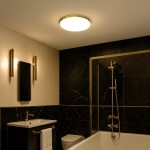 Plafon Maya LED D33 o średnicy 33 cm w kolorze złotym, z białym kloszem, zamontowany na suficie w nowoczesnej łazience. Oświetlenie ma klasę IP44, moc 15W i barwę 4000K, idealne do strefy mokrej. Otoczenie w ciemnych tonach z elementami złota.
