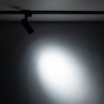 Reflektor spot Tracer 1xLED 10W w kolorze czarnym, zamontowany na szynie 1-fazowej. Ujęcie z boku, oświetlający jasno obszar na ścianie, z widoczną strukturą materiału i światłem w kształcie elipsy.