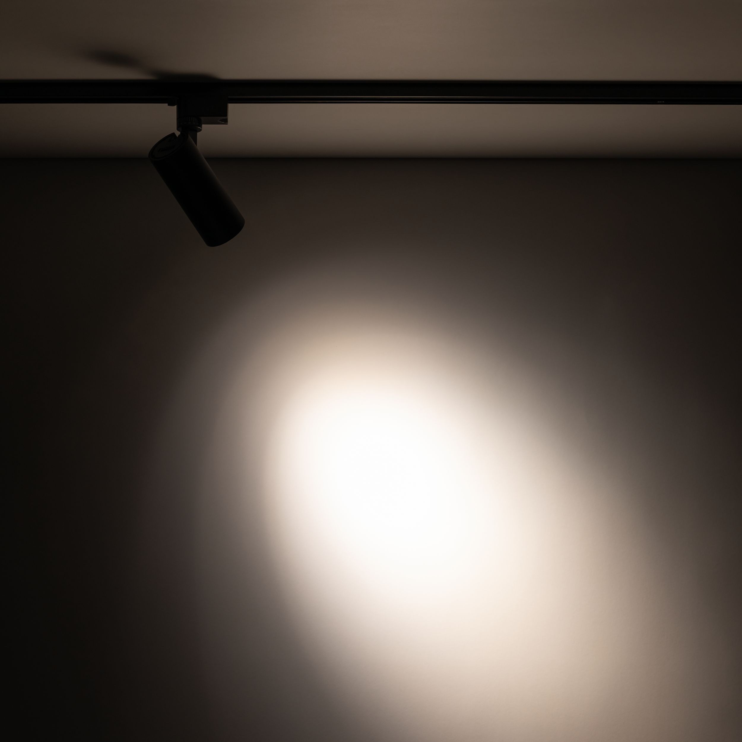 Reflektor Tracer 1xLED 10W CCT w kolorze czarnym, zamontowany na szynie 1-fazowej. Ujęcie z boku, ukazujące źródło światła i plamę światła na ścianie w tle. Produkt o cylindrycznym kształcie, wykonany z metalu.