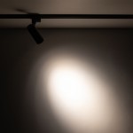 Reflektor Tracer 1xLED 10W CCT w kolorze czarnym, zamontowany na szynie 1-fazowej. Ujęcie z boku, ukazujące źródło światła i plamę światła na ścianie w tle. Produkt o cylindrycznym kształcie, wykonany z metalu.