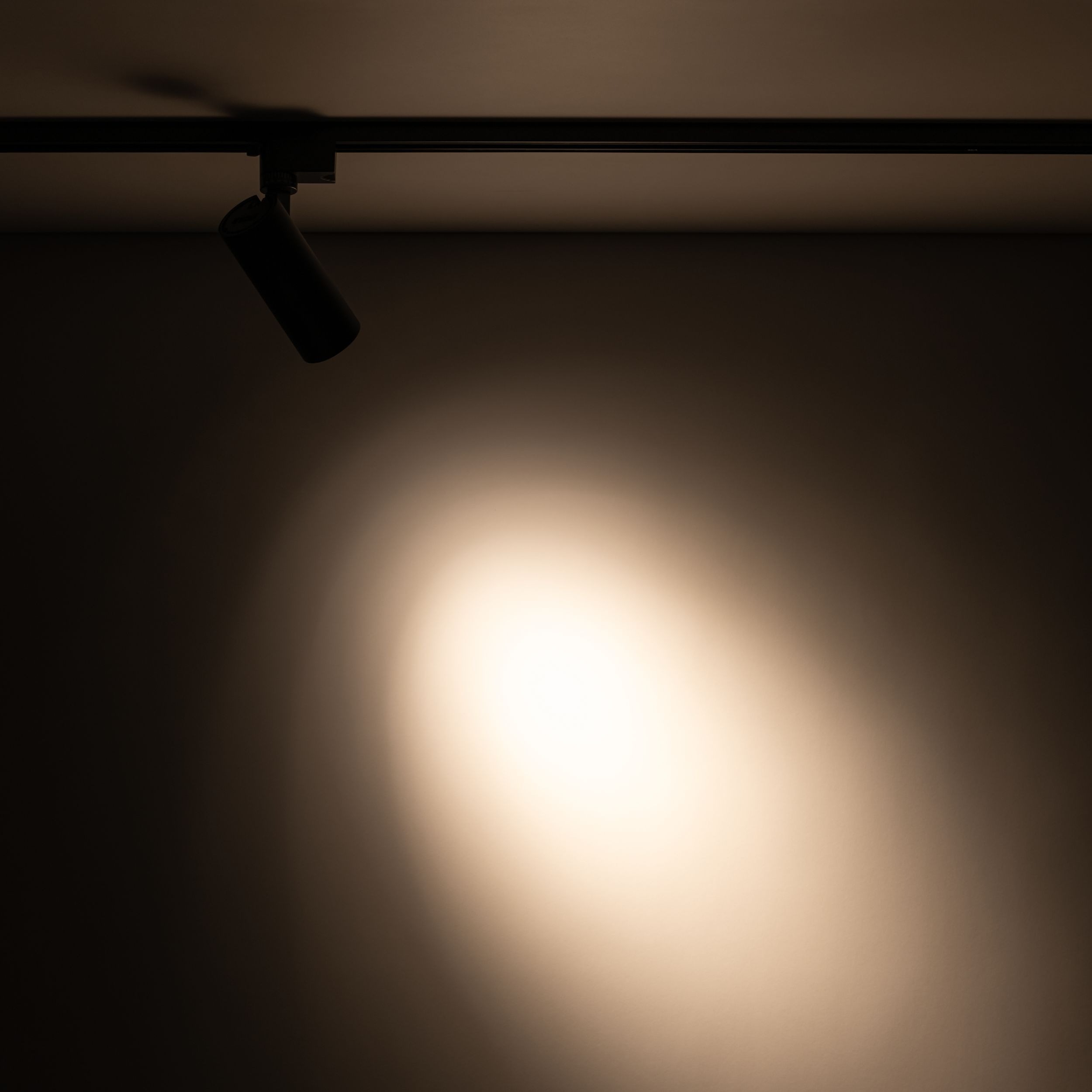 Reflektor spot Tracer 1xLED 10W CCT w kolorze czarnym, zamontowany na szynie 1-fazowej, skierowany w dół, emitujący ciepłe światło na ścianę, z wyraźnym punktem świetlnym w tle i delikatnym przejściem do ciemniejszego otoczenia.