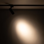 Reflektor spot Tracer 1xLED 10W CCT w kolorze czarnym, zamontowany na szynie 1-fazowej, skierowany w dół, emitujący ciepłe światło na ścianę, z wyraźnym punktem świetlnym w tle i delikatnym przejściem do ciemniejszego otoczenia.