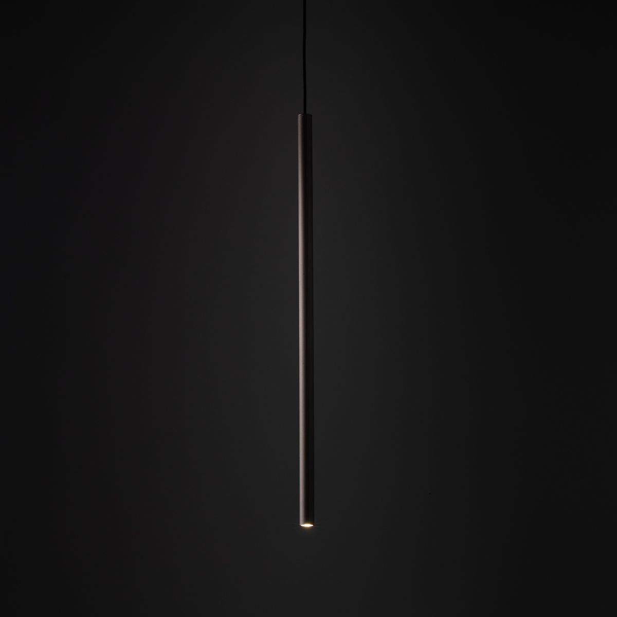 Brązowa lampa wisząca Tracer Brown 1xG9 do szyny, w formie długiego, prostokątnego tubusu. Ujęcie frontalne, lampa zawieszona na cienkim przewodzie, emitująca delikatne światło z dolnej części. Tło ciemne, podkreślające elegancki design.