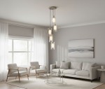 Lampa wisząca Duo I, 5-punktowa, w srebrnej satynie i przezroczystym kloszu. Umieszczona w nowoczesnym wnętrzu, w tle widoczne okno z zasłonami oraz jasna sofa z poduszkami. Lampa zawieszona na długich przewodach, tworzy elegancką aranżację.