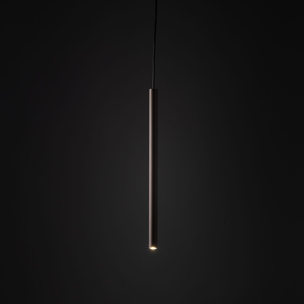 Lampa wisząca Tracer 1xG9, brązowa, w kształcie smukłego, cylindrycznego słupka. Zawieszona na cienkim przewodzie, emitująca światło z dolnej części. Tło ciemne, co podkreśla minimalistyczny design lampy. Idealna do szyn 1-fazowych.