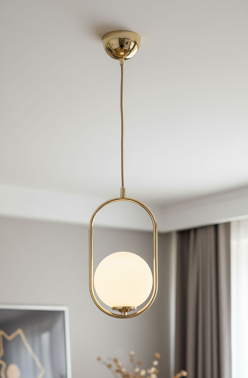Lampa wisząca Noval M 1-punktowa o złotej metalowej obudowie z kulistym, mlecznym kloszem. Produkt zawieszony na cienkim kablu, umieszczony w nowoczesnym wnętrzu z szarym tłem i zasłonami.