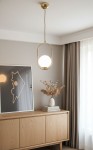 Lampa wisząca Noval M w kolorze złotym, 1-punktowa, z okrągłym, białym kloszem. Zawieszona na cienkim przewodzie, w aranżacji wnętrza z drewnianym meblem i dekoracjami. Tło w neutralnych odcieniach.
