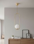 Lampa wisząca Noval L 1-punktowa w złotym wykończeniu z białym, okrągłym kloszem. Ujęcie frontalne, zawieszona na długim przewodzie. Tło jasnoszare z drewnianą komodą oraz dekoracjami, takimi jak roślina i ramki ze zdjęciami.