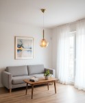 Lampa wisząca Grall D20 w złotym kolorze z przezroczystym kloszem, zawieszona na cienkim kablu w jasnym wnętrzu. W tle szara sofa, drewniany stół z rośliną i książkami, oraz okno z białymi firanami.