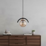 Złota lampa wisząca Korda 1-punktowa z przezroczystą kulistą kloszem, zawieszona na czarnym przewodzie. Kształt owalny z metalowym wykończeniem. Tło w odcieniach szarości, drewniana komoda z dekoracjami w dolnej części.
