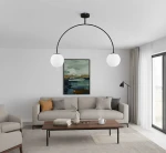 Czarna lampa wisząca Bendo z dwoma okrągłymi kloszami w białym kolorze, zamontowana na czarnym stelażu w kształcie łuku. Widoczna w nowoczesnym wnętrzu z szaro-beżową sofą i drewnianym stolikiem.