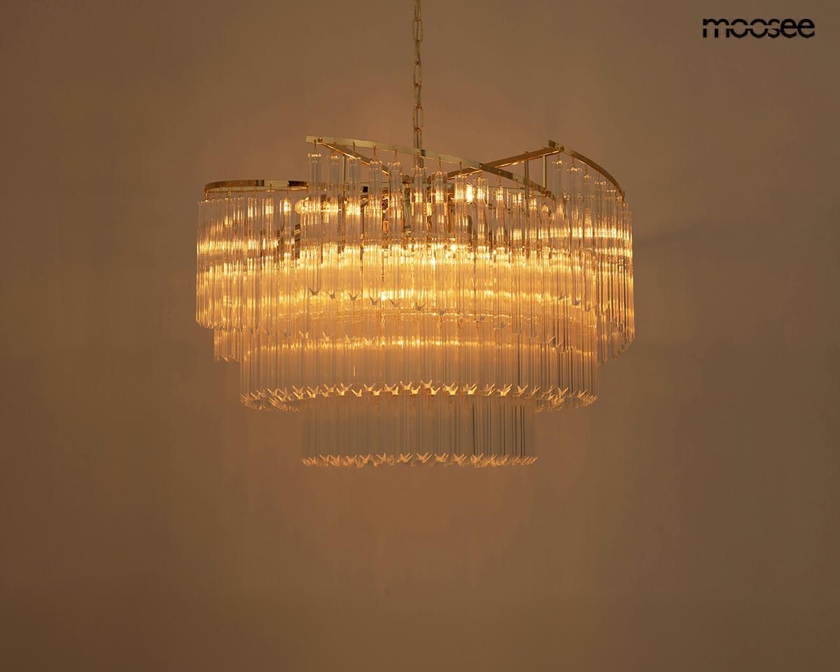 Lampa wisząca Luma 60 w kolorze złotym, o konstrukcji z przezroczystych elementów. Posiada dziewięć miejsc na żarówki E14, zawieszona na łańcuchu, w górnej części o zaokrąglonym kształcie, emitująca ciepłe światło. Tło proste.