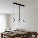 Lampa wisząca Sitro w kolorze czarnym i białym, z trzema okrągłymi kloszami z mlecznego szkła zawieszonymi na długich linkach. Ujęcie frontalne, lampy zamontowane na prostokątnej, czarnej listwie, w tle stół z nakryciem w nowoczesnym wnętrzu.