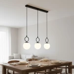 Lampa wisząca Sitro z trzema kulistymi kloszami w białym kolorze, zawieszona na czarnej listwie. Produkt umieszczony nad drewnianym stołem w jasnym wnętrzu, idealny do jadalni. Klosze zawieszone na cienkich linkach.