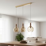 Lampa wisząca Hetty 3xE27 w kolorze złotym z drewnianymi detalami. Składa się z trzech szklanych kloszy w kształcie kul, zawieszonych na cienkich linkach, montowanych do prostokątnej podstawy. Tło to jasne wnętrze z rośliną i stołem.