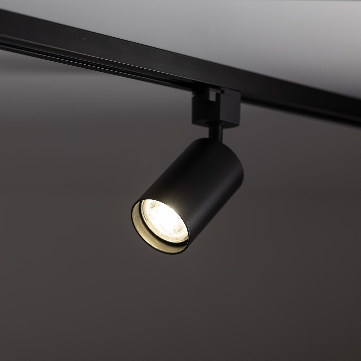 Reflektor spot Tracer 1xGU10 w kolorze czarnym, zamontowany na szynie 1-fazowej. Cylindryczny kształt z widoczną żarówką emitującą światło. Ujęcie z boku, ukazujące detal i wykończenie lampy. Tło proste, ciemne.