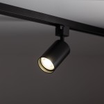 Reflektor spot Tracer 1xGU10 w kolorze czarnym, zamontowany na szynie 1-fazowej. Cylindryczny kształt z widoczną żarówką emitującą światło. Ujęcie z boku, ukazujące detal i wykończenie lampy. Tło proste, ciemne.