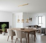 Lampa wisząca Volda 3-punktowa w kolorze złotym z okrągłymi kloszami, zawieszona na prostokątnej podstawie, umieszczona nad drewnianym stołem w nowoczesnej jadalni. W tle widoczna roślina i kanapa.