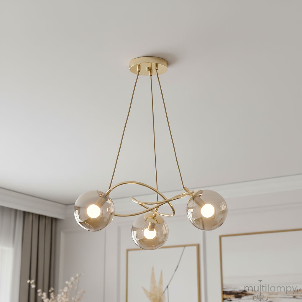 Lampa wisząca Velta 3-punktowa w kolorze złotym z trzema przezroczystymi kulistymi kloszami, zawieszona na cienkich linkach. Klosze mają delikatne, białe źródła światła. Produkt umieszczony w minimalistycznym wnętrzu.