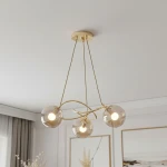 Lampa wisząca Velta 3-punktowa w kolorze złotym z trzema przezroczystymi kulistymi kloszami, zawieszona na cienkich linkach. Klosze mają delikatne, białe źródła światła. Produkt umieszczony w minimalistycznym wnętrzu.