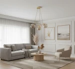 Lampa wisząca Velta 3-punktowa w kolorze złotym, z trzema szklanymi kloszami, zawieszona na smukłych linkach. Produkt umieszczony w eleganckim wnętrzu z jasnymi ścianami, sofą i drewnianym stolikiem.