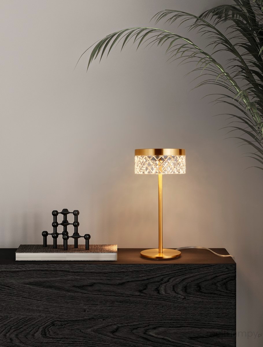 Lampa stołowa Mineros 1xLED 6W w kolorze złotym, z przezroczystym kloszem o wzorze diamentowym. Ustawiona na ciemnym blacie, obok książek i dekoracji. Kształt cylindryczny, z cienką, prostą podstawą.