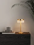 Lampa stołowa Mineros 1xLED 6W w kolorze złotym, z przezroczystym kloszem o wzorze diamentowym. Ustawiona na ciemnym blacie, obok książek i dekoracji. Kształt cylindryczny, z cienką, prostą podstawą.