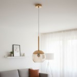 Lampa wisząca Volda 1-punktowa w kolorze złotym, z przezroczystym kloszem w formie półkuli. Zawieszona na białym przewodzie, montowana do sufitu. W tle widoczny stół z rośliną oraz miękką kanapą.