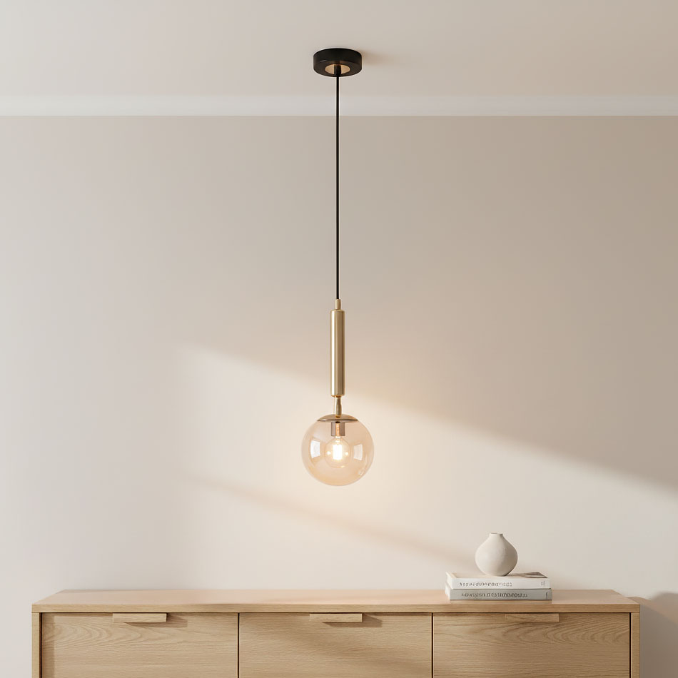 Czarna lampa wisząca Volda Picolo z złotym detalem, zawieszona na cienkim kablu. Szklany klosz w kształcie kuli, przezroczysty, z widoczną żarówką. Produkt umieszczony nad drewnianym meblem z jasnego drewna, na tle jasnej ściany.