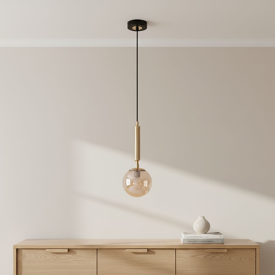 Lampa wisząca Volda Picolo 1-punktowa w kolorze czarnym i złotym, z przezroczystym kloszem w kształcie kuli. Zawieszona na cienkim, czarnym przewodzie, montowana do sufitu. W tle drewniana komoda z dekoracjami.