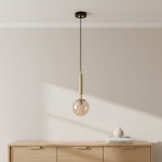 Lampa wisząca Volda Picolo 1-punktowa w kolorze czarnym i złotym, z przezroczystym kloszem w kształcie kuli. Zawieszona na cienkim, czarnym przewodzie, montowana do sufitu. W tle drewniana komoda z dekoracjami.