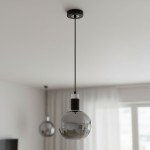 Lampa wisząca Adel z jedną oprawą E27, w kolorze czarnym z chromowanymi detalami. Okrągła, szklana kula w odcieniu szaro-przezroczystym, zawieszona na cienkim, czarnym przewodzie w minimalistycznym wnętrzu.