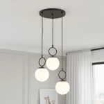 Lampa wisząca Sitro z trzema kulistymi kloszami w białym kolorze, zawieszona na czarnych linkach, z okrągłymi elementami mocującymi. Zainstalowana na sufitowej podstawie w kolorze czarnym, w minimalistycznym wnętrzu.