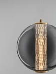 Lampa wisząca Morros 1xLED 8W w kolorze złotym. Okrągła, przezroczysta kula z szarego szkła, z centralnym, teksturowanym elementem w odcieniach złota. Zawieszona na cienkim kablu, idealna do nowoczesnych wnętrz.