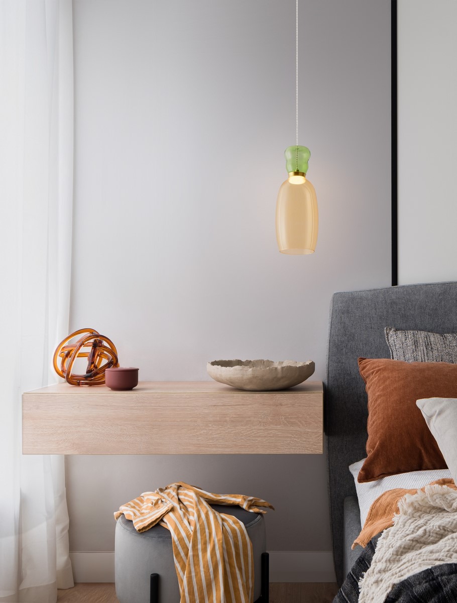 Lampa wisząca Perene 1xLED 9W w kolorze złotym i żółtym, z przezroczystym kloszem, zawieszona nad drewnianym stolikiem nocnym. W tle widoczne poduszki w odcieniach brązu i szarości oraz dekoracyjne akcesoria.